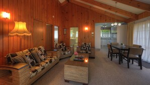 A cozy living room with a sofa, armchairs, and a coffee table. There is a dining area with a table and chairs. The room has wooden walls and a high ceiling. - Shiralee Executive Cottages (Norfolk Island)