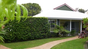 A house with a paved driveway leading to a front door, surrounded by a well-maintained lawn and a tall hedge. - Shiralee Executive Cottages (Norfolk Island)
