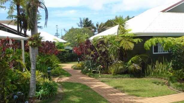 A garden pathway leading to a building with a white roof, surrounded by lush tropical plants and palm trees. - Shiralee Executive Cottages (Norfolk Island)