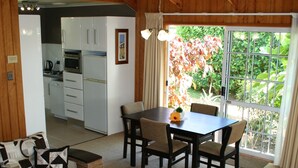 A cozy living area with a sofa, a coffee table, and a dining table with chairs. There is a kitchen in the background with white cabinets and a refrigerator. - Shiralee Executive Cottages (Norfolk Island)