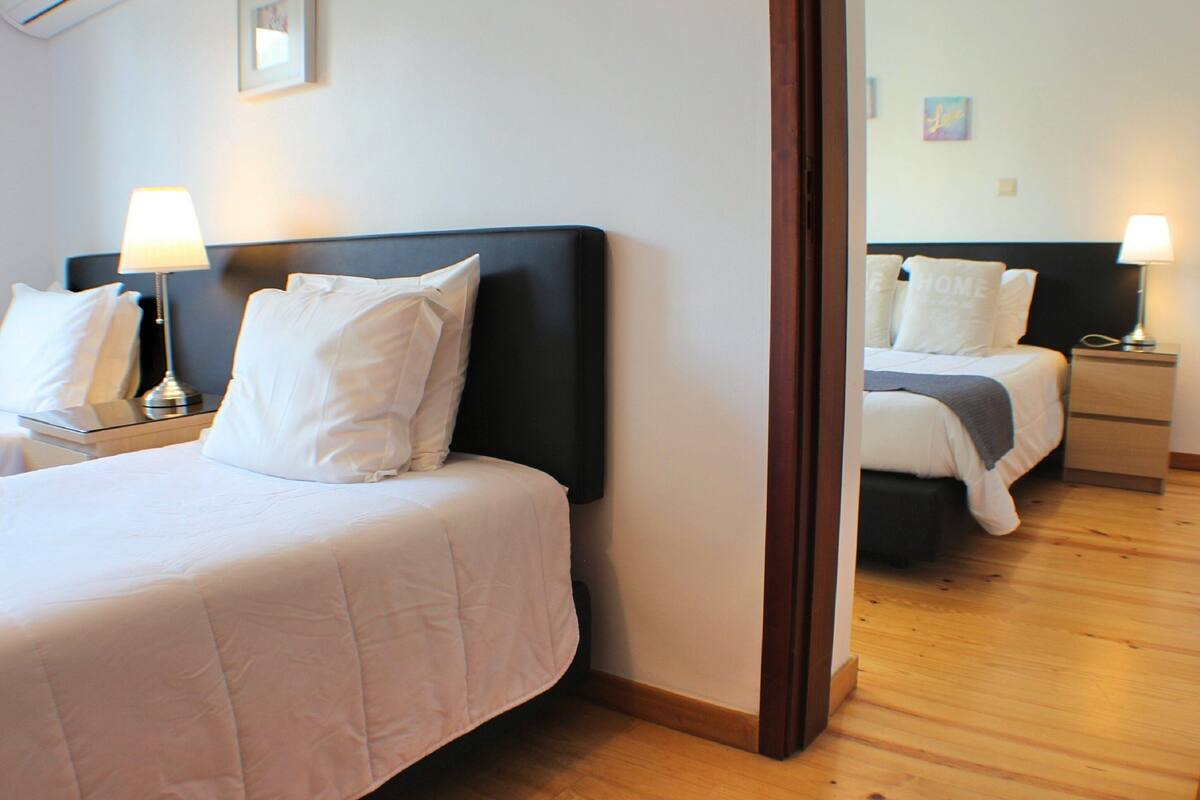A hotel room with two beds, each with white bedding and a dark headboard. There are bedside tables with lamps and a small picture frame on the wall.