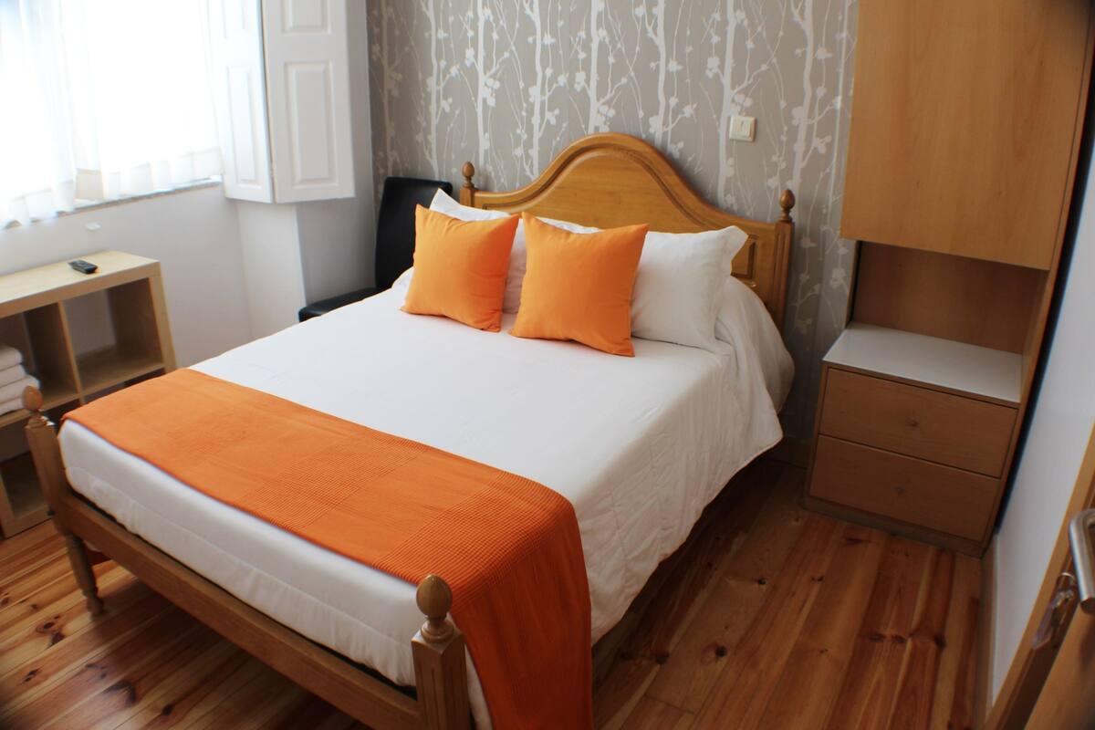 A neatly made bed with an orange blanket and pillows, a wooden headboard, a bedside table, and a nightstand.