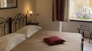 A bedroom with a bed, a nightstand, a framed picture, a window with curtains, and a view of buildings outside.