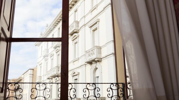 A view through a window with a metal balcony railing, looking at a white building with classical architecture.