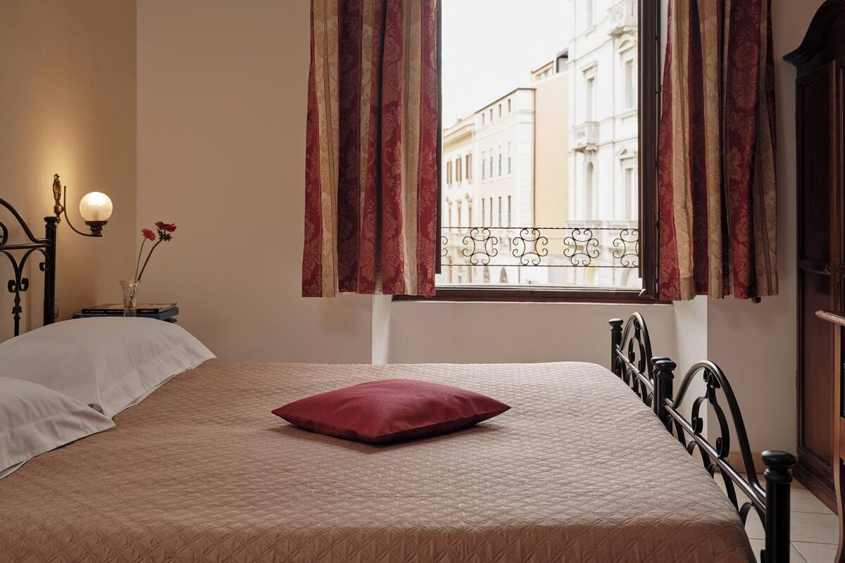 A bedroom with a bed, a nightstand with a lamp, a vase with flowers, and a window with curtains.