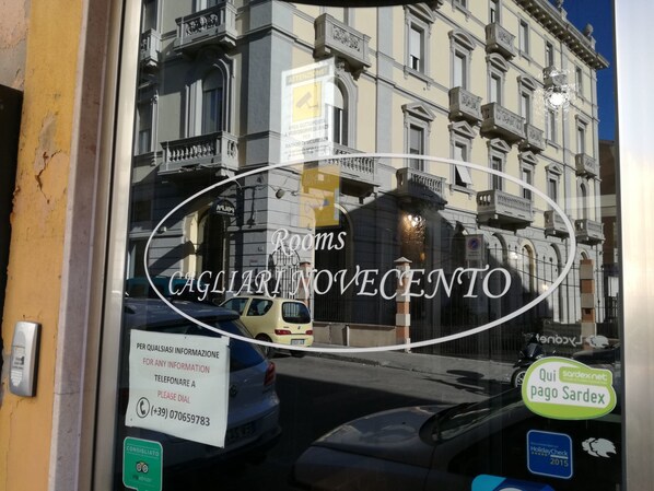 Janela de hotel com um letreiro indicando 'Rooms CAGLIARINO VECENTO' e um número de contato.