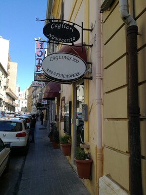 Rua estreita com um hotel identificado pelas placas "Cagliari 900" e "Cagliari Wavecento".