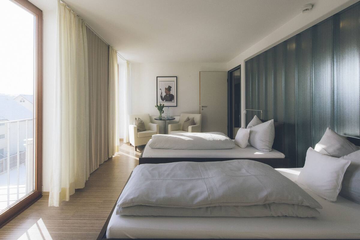 A hotel room with two beds, a sitting area with a table and chairs, a framed picture on the wall, and a large window offering a view.