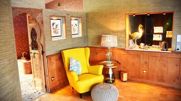 A room with a wooden floor, a yellow armchair, a round ottoman, a lamp, framed artwork on the wall, and a wooden cabinet with a display.