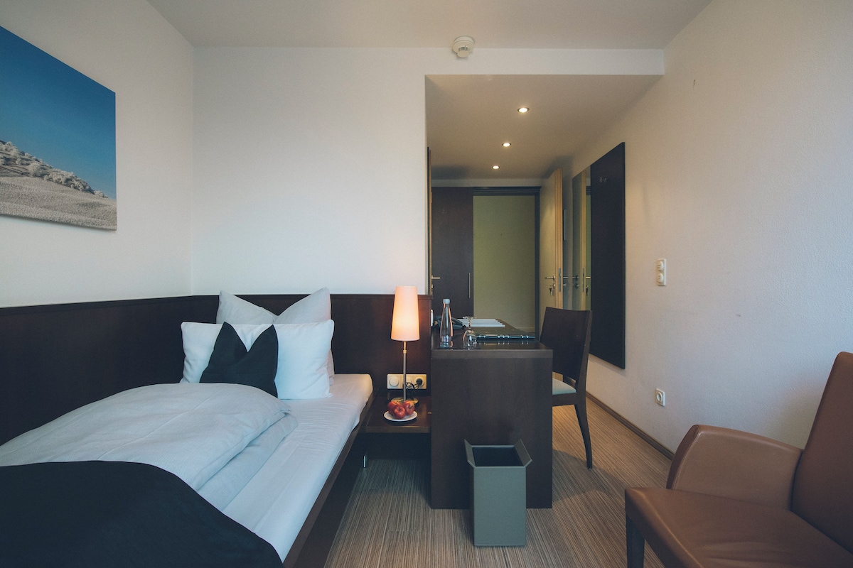 A hotel room with a bed, a desk, a chair, and a small trash bin.