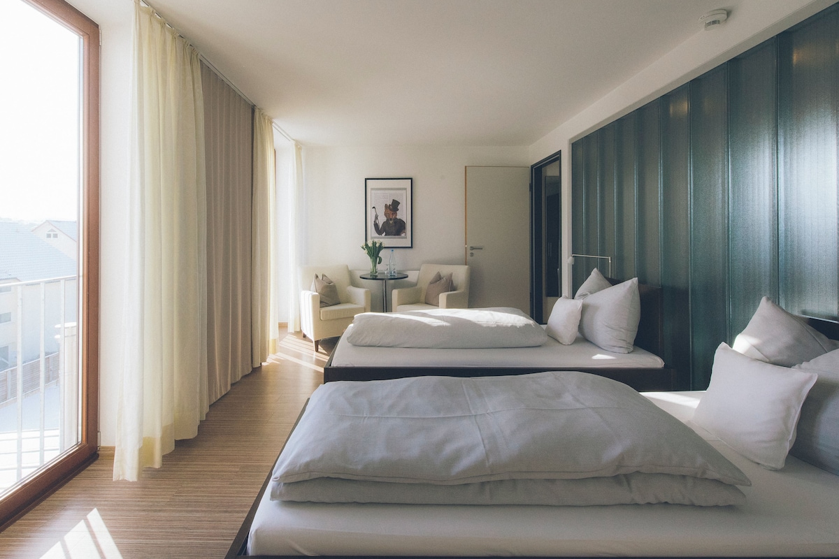 A hotel room with two beds, a sitting area with a table and chairs, a framed picture on the wall, and a large window offering a view.