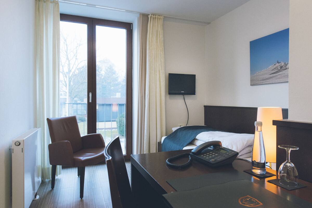 A hotel room with a bed, a chair, a desk with a phone and a bottle, a TV, and a window with curtains.
