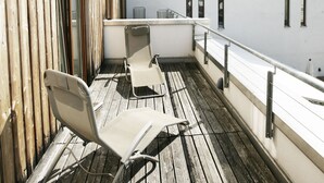 Een houten terras met twee lounge fauteuils en een kleine tafel, met uitzicht op een gebouw met grote ramen en een witte gevel.