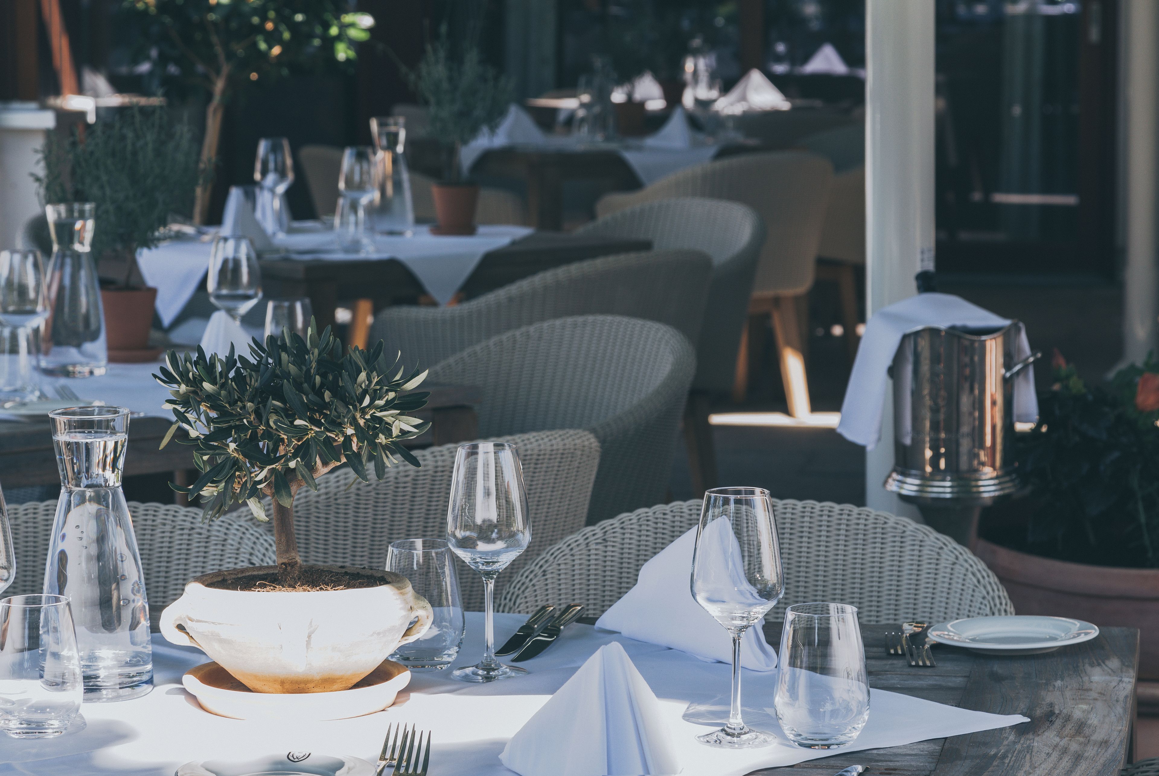 Un área de comedor al aire libre con mesas dispuestas para dos, con copas transparentes, una planta en maceta y sillas de mimbre.