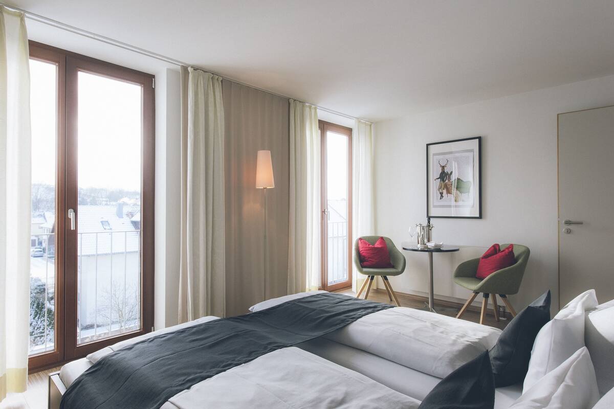 A hotel room with a bed, a chair, a small table, and a window with curtains.