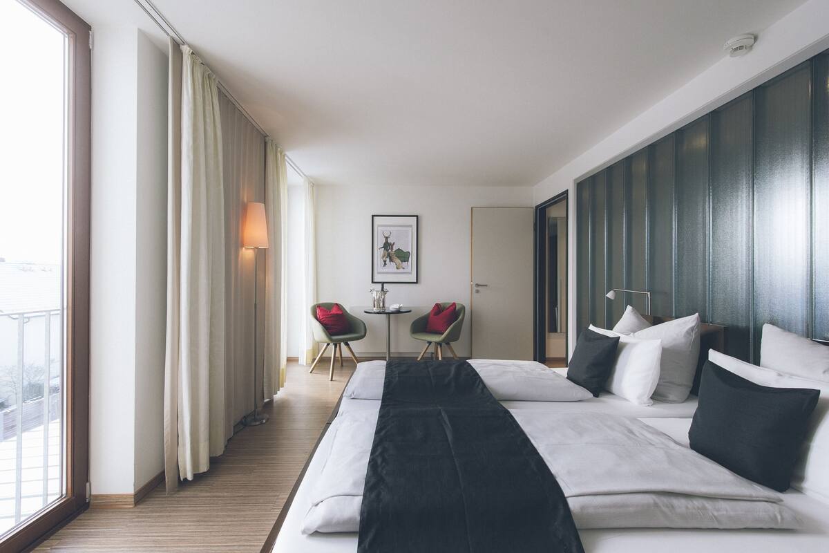 A modern hotel room with a large bed, two chairs, a small table, and a view of the outdoors.