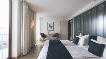 A modern hotel room with a large bed, two chairs, a small table, and a view of the outdoors.