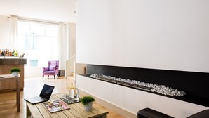A modern living room with a wooden coffee table, a laptop, a magazine, and a potted plant. There is a fireplace with white pebbles and a purple armchair. - Hôtel Atrium Mondial (Lourdes)