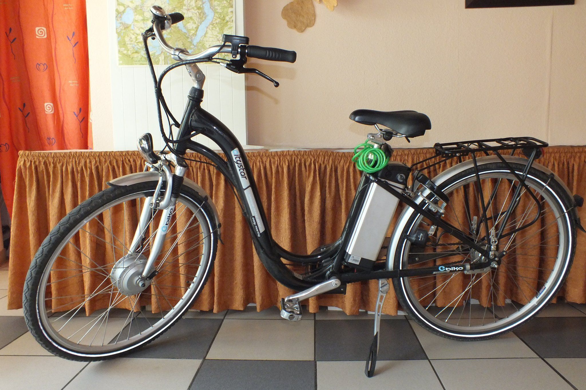 A stationary bicycle with a seat, handlebars, and a rear rack, positioned against a wall with a curtain and a map.