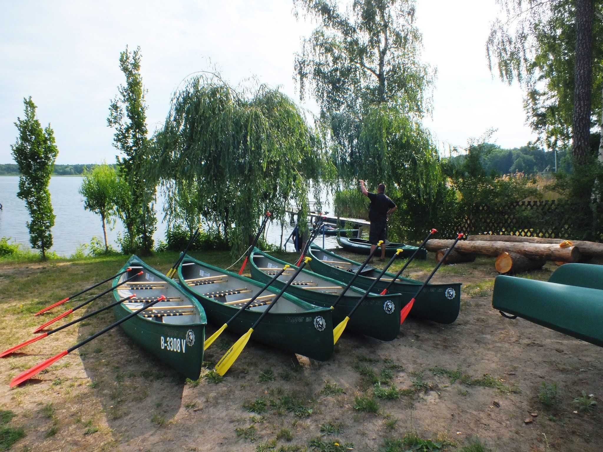 Several green canoes lined up on a grassy area near a lake, with a person standing nearby.
