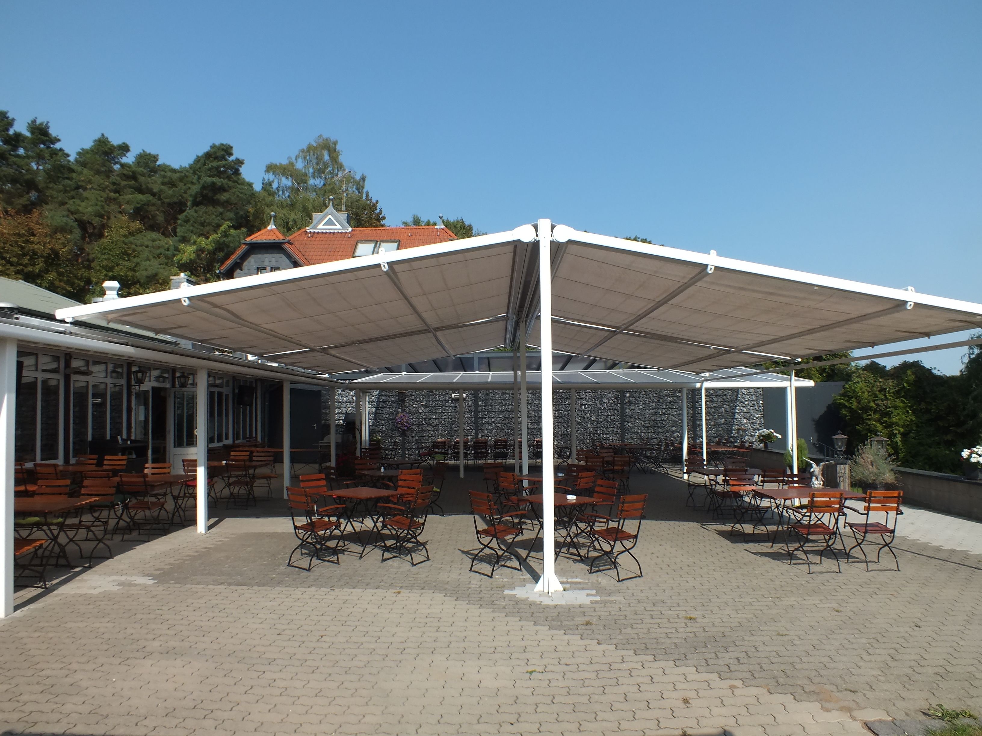 Um pátio externo com mesas e cadeiras, coberto por um toldo branco, e uma parede de pedra ao fundo.