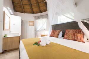Habitación de hotel con cama, mesita de noche, espejo, ventana con cortinas y techo de paja.