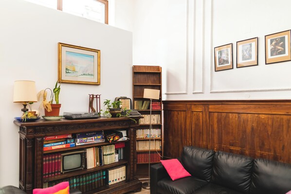 A cozy living room with a leather sofa, a bookshelf filled with books and board games, a small table with a lamp and a potted plant, and a wooden cabinet. - Ostello Domus Civica (Venice)