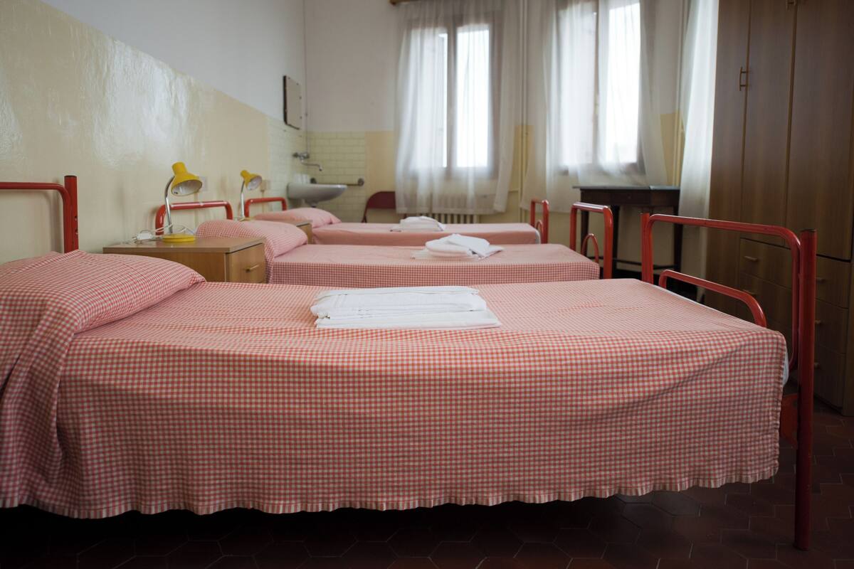A hotel room with two single beds, each with a checkered bedspread and neatly folded towels. There is a small bedside table with a lamp, a window with curtains, and a radiator.