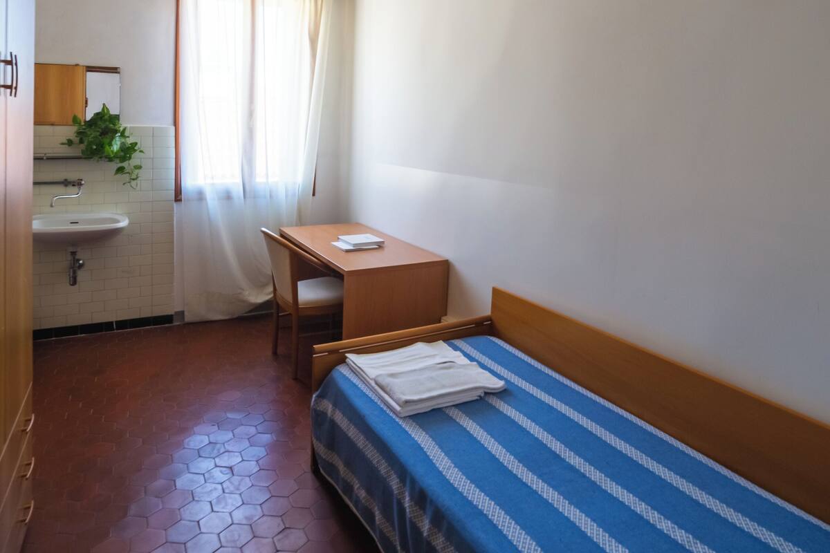 A small, compact room with a single bed, a wooden desk, a chair, a small table, a sink, and a mirror.