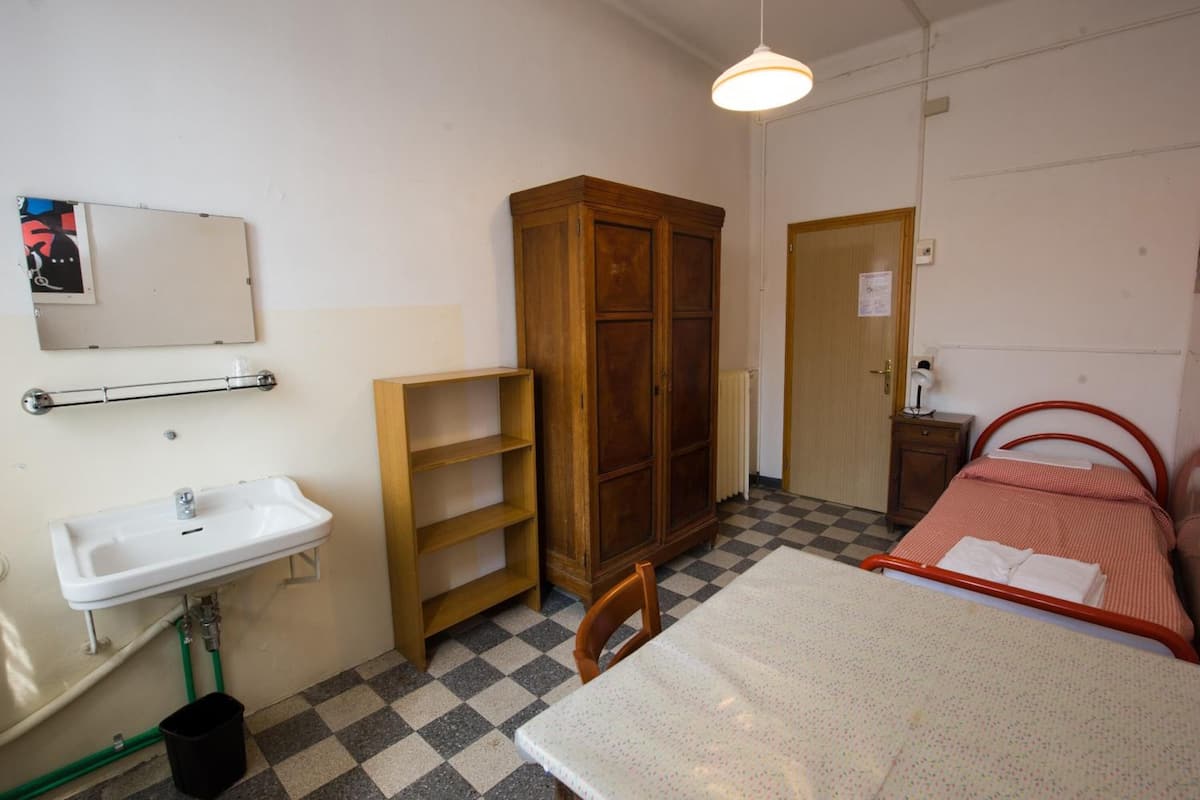 A small, compact room with a single bed, a wooden wardrobe, a white sink, and a mirror on the wall.