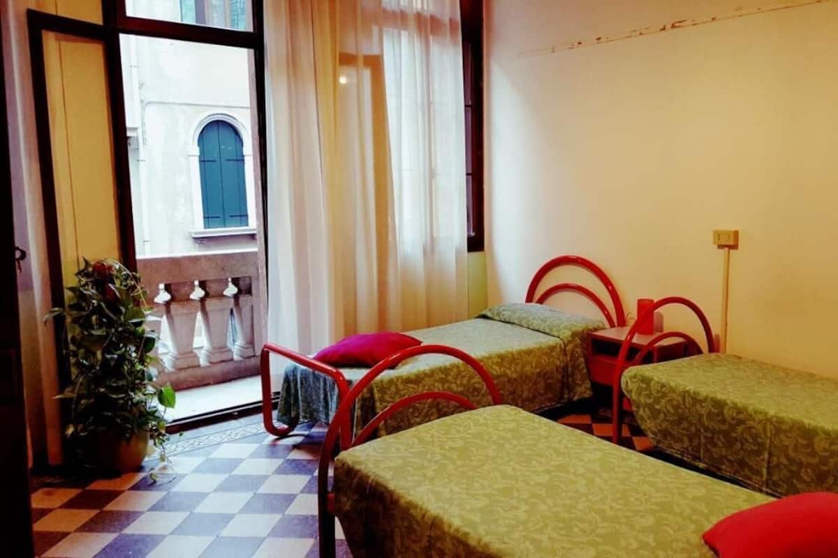 A room with two single beds, a small table, and chairs. There is a potted plant and a view of a building through a window.