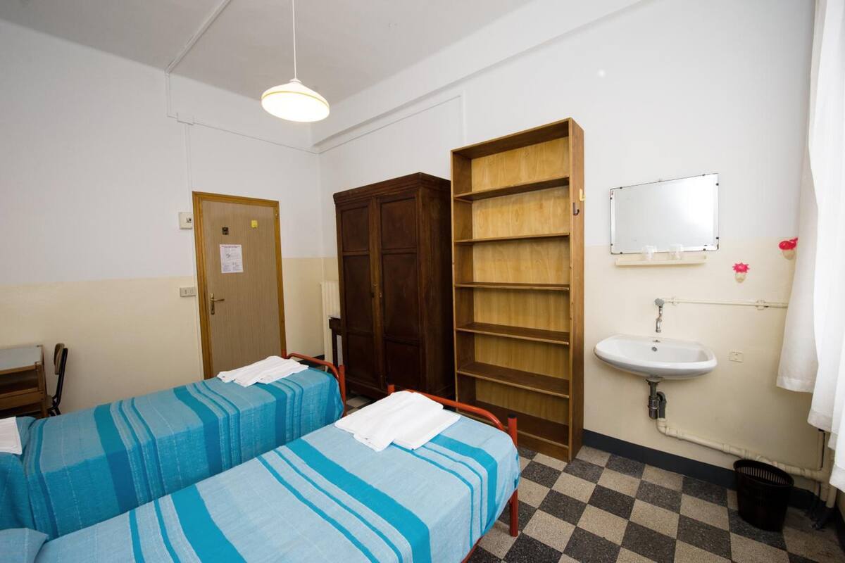 A room with two beds, a wooden wardrobe, a shelf unit, a sink, and a mirror.