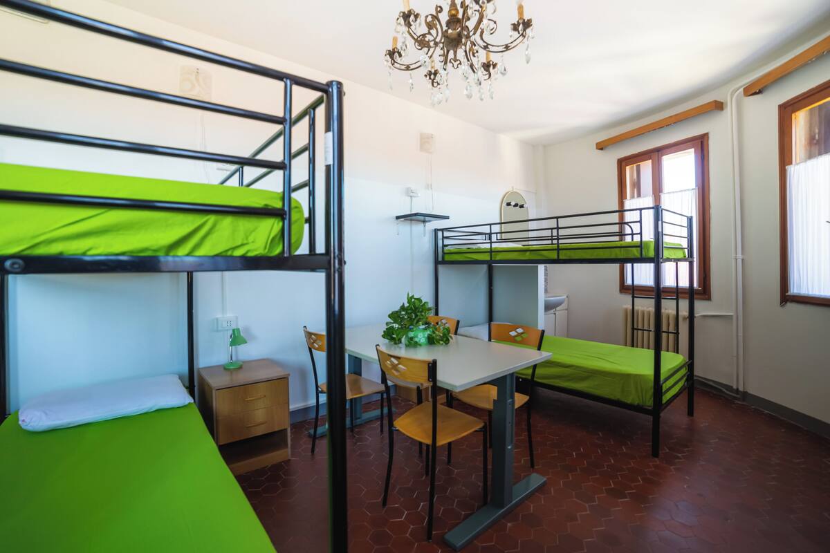 A dormitory room with bunk beds, a dining table, chairs, a small desk, and a chandelier.
