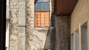 A narrow alleyway with two stone buildings, one with a wooden door and window shutters, and the other with a wooden door and a window with a pointed arch.