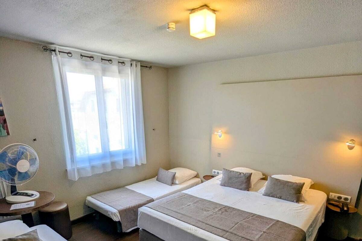 A hotel room with two single beds, a double bed, a standing fan, and a window with curtains.