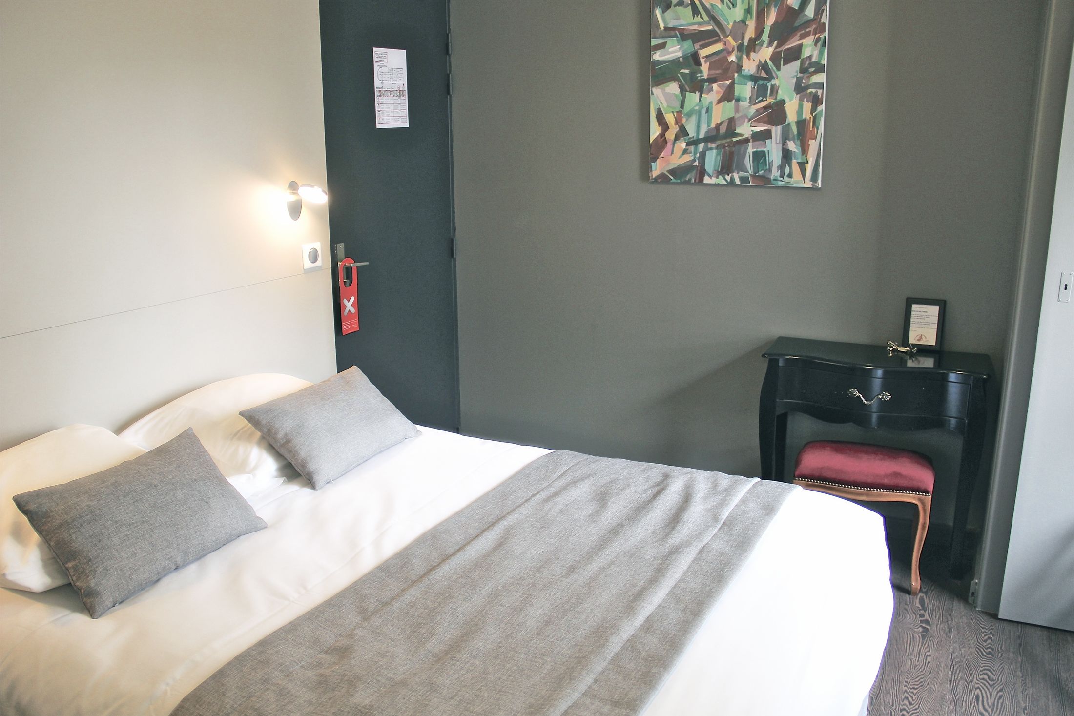 Een netjes opgemaakt bed met een grijs-witte spreideken, een klein rode gestoffeerde bankje, een nachtkastje van donker hout met een lade, en een wandlamp.