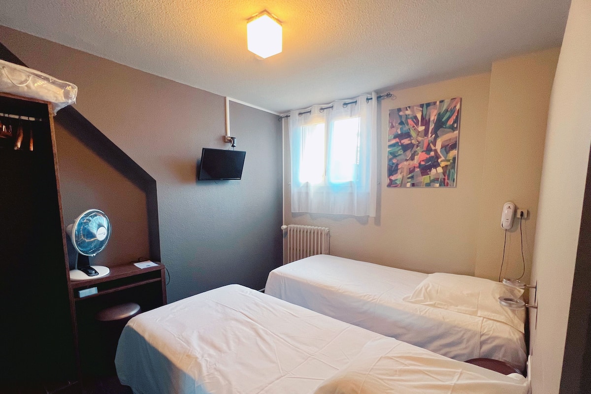 A hotel room with two beds, a desk with a fan, a TV, a radiator, and a painting on the wall.