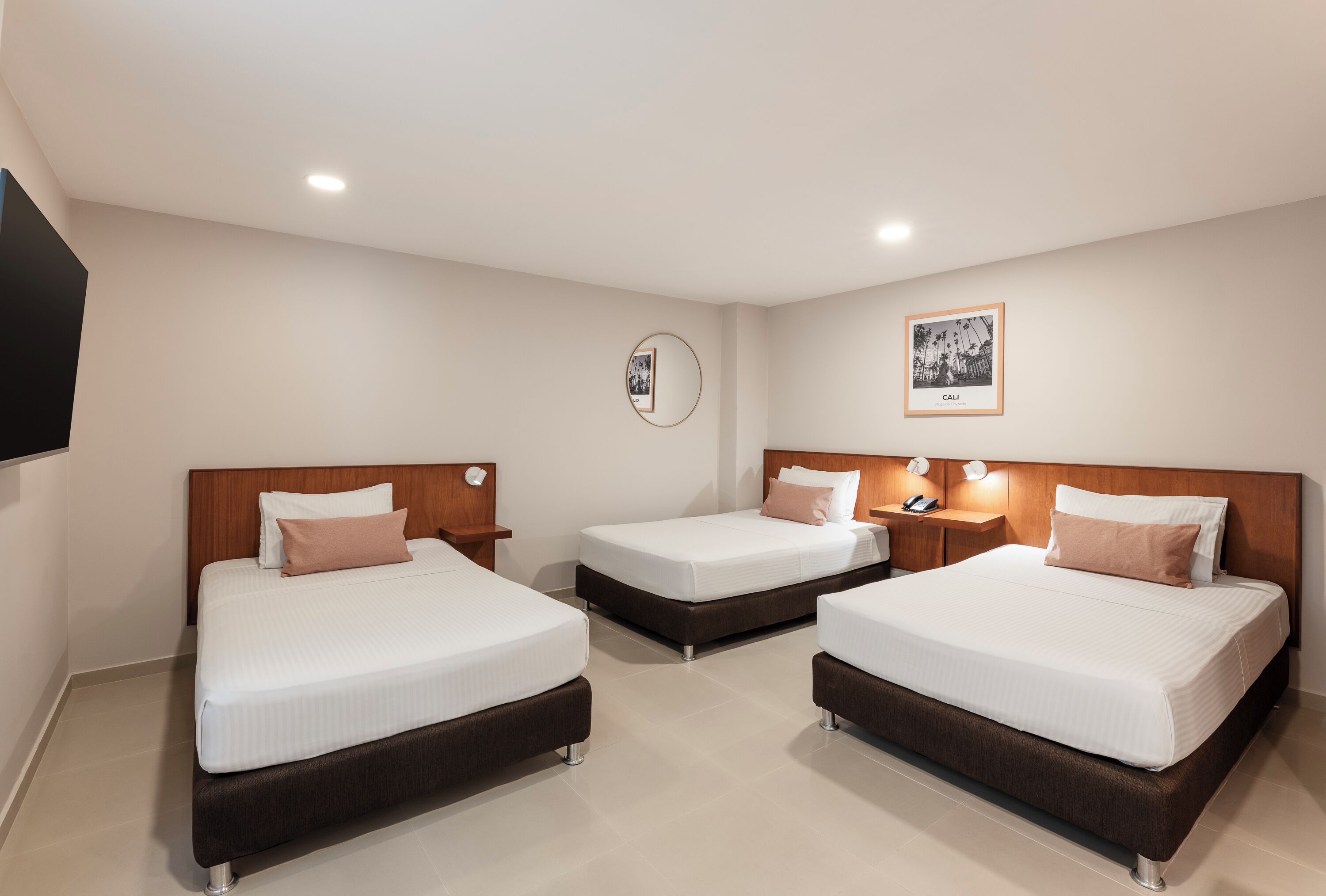 A hotel room with three beds, a round mirror, a framed picture on the wall, and a bedside table with a lamp.