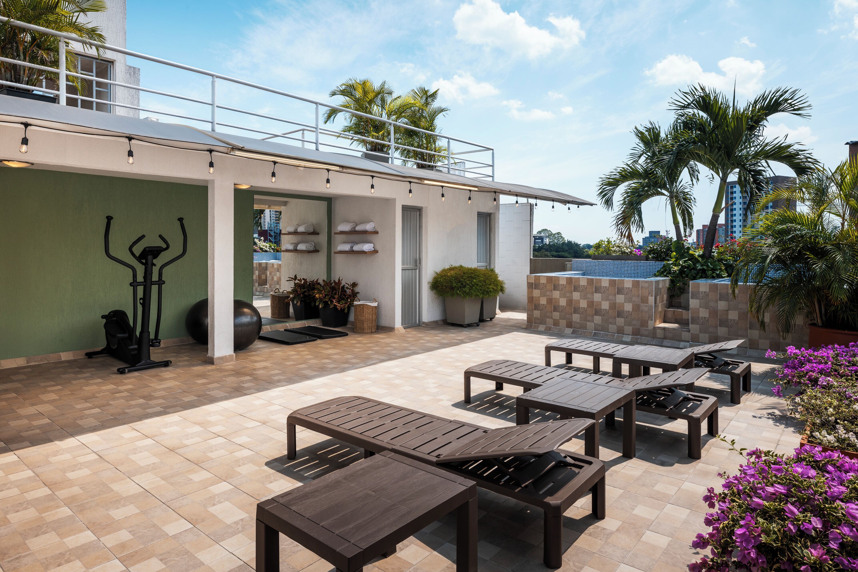 Un espace extérieur aménagé en terrasse, avec plusieurs tables en bois et des fauteuils de jardin, un espace dédié à la remise en forme avec des appareils d’exercice, et une vue sur des palmiers et des bâtiments en arrière-plan.
