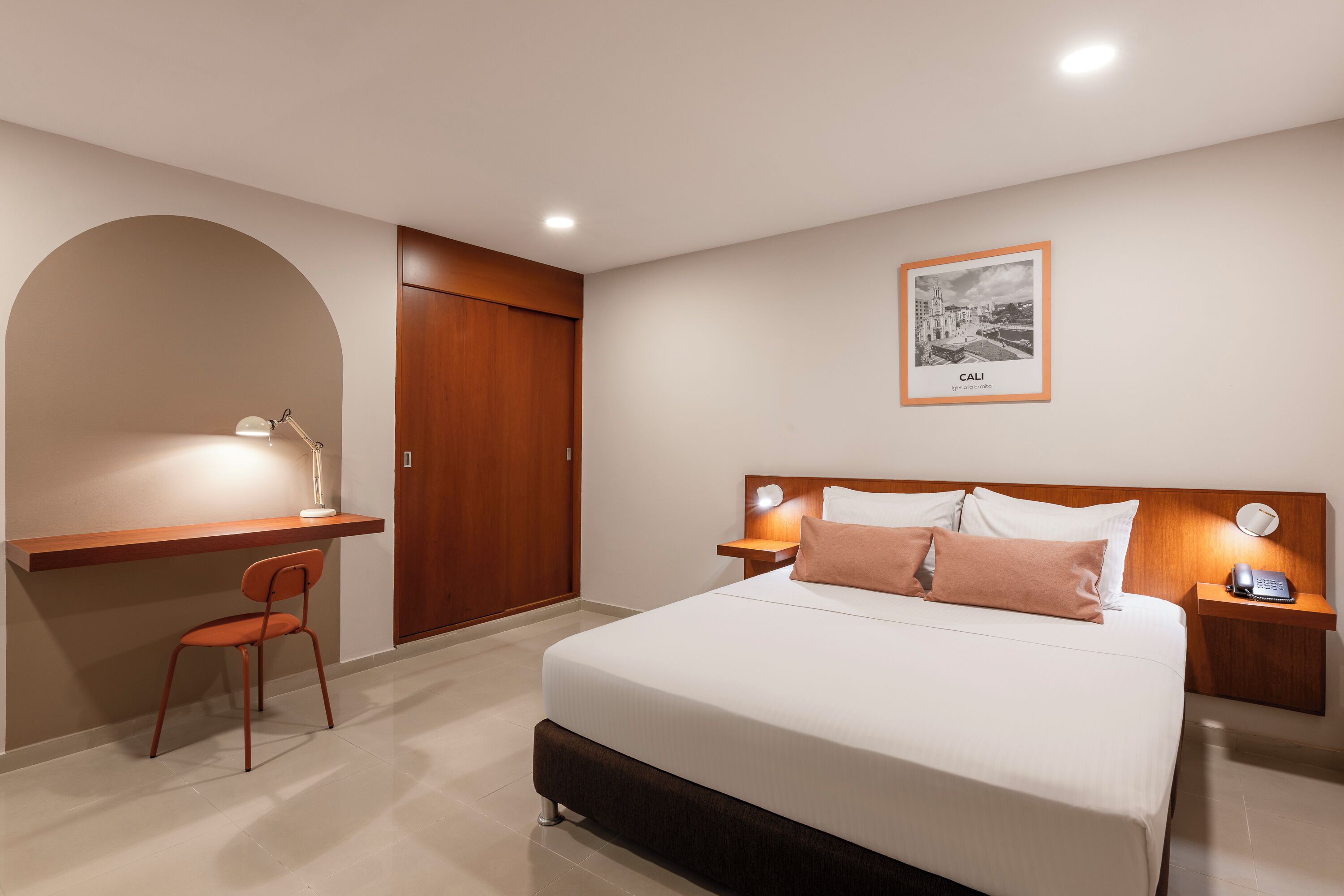 A hotel room with a bed, bedside tables, a desk with a chair, a wardrobe, and a framed picture on the wall.