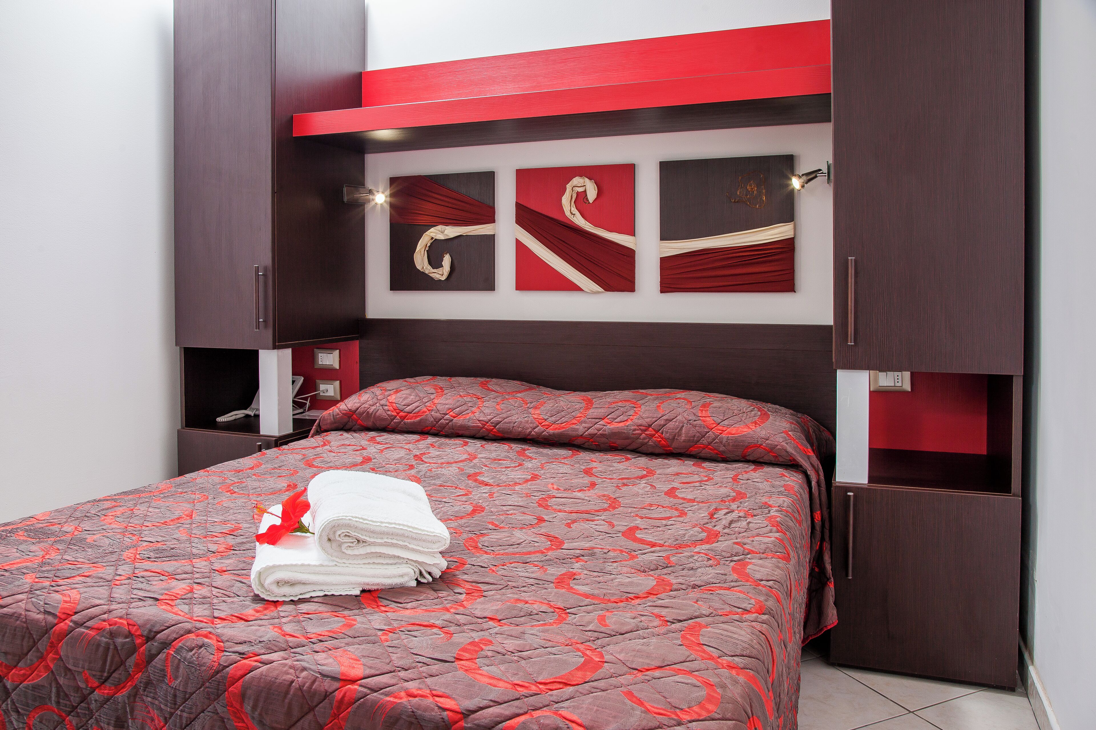 A neatly made bed with a red and brown patterned bedspread, a folded white towel, and a red rose. There are three wall-mounted artworks above the bed, and a small shelf with a lamp to the left.