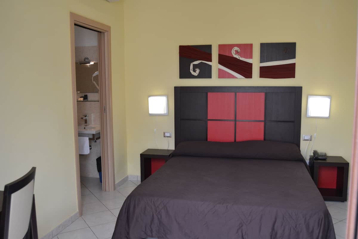 A bedroom with a large bed, two bedside tables, a nightstand, a wall-mounted TV, and a door leading to a bathroom.
