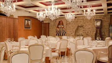 A formal dining room with multiple tables and chairs, chandeliers, and a clock on the wall.