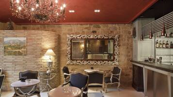 A bar area with a stone wall, a mirror, and a chandelier. There are tables and chairs for seating.