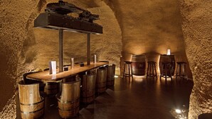 Une cave à vin souterraine, avec des barriques en bois, une longue table et un porte-bouteilles suspendu.