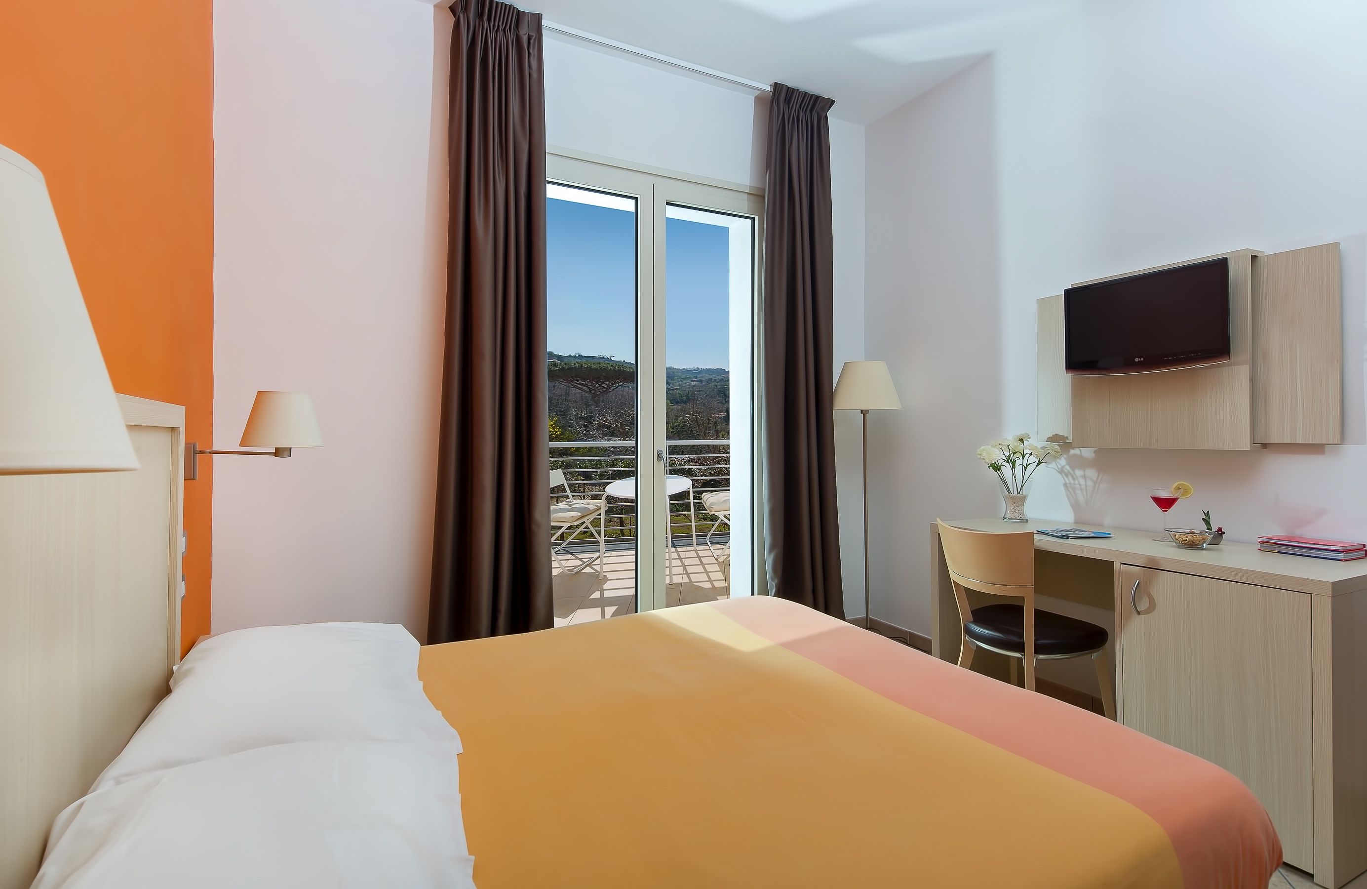 A hotel room with a bed, a desk with a chair, a TV, and a balcony with chairs.