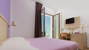 Een hotelkamer met een bed, een bureau met een televisie, een raam met gordijnen en uitzicht op de lucht.