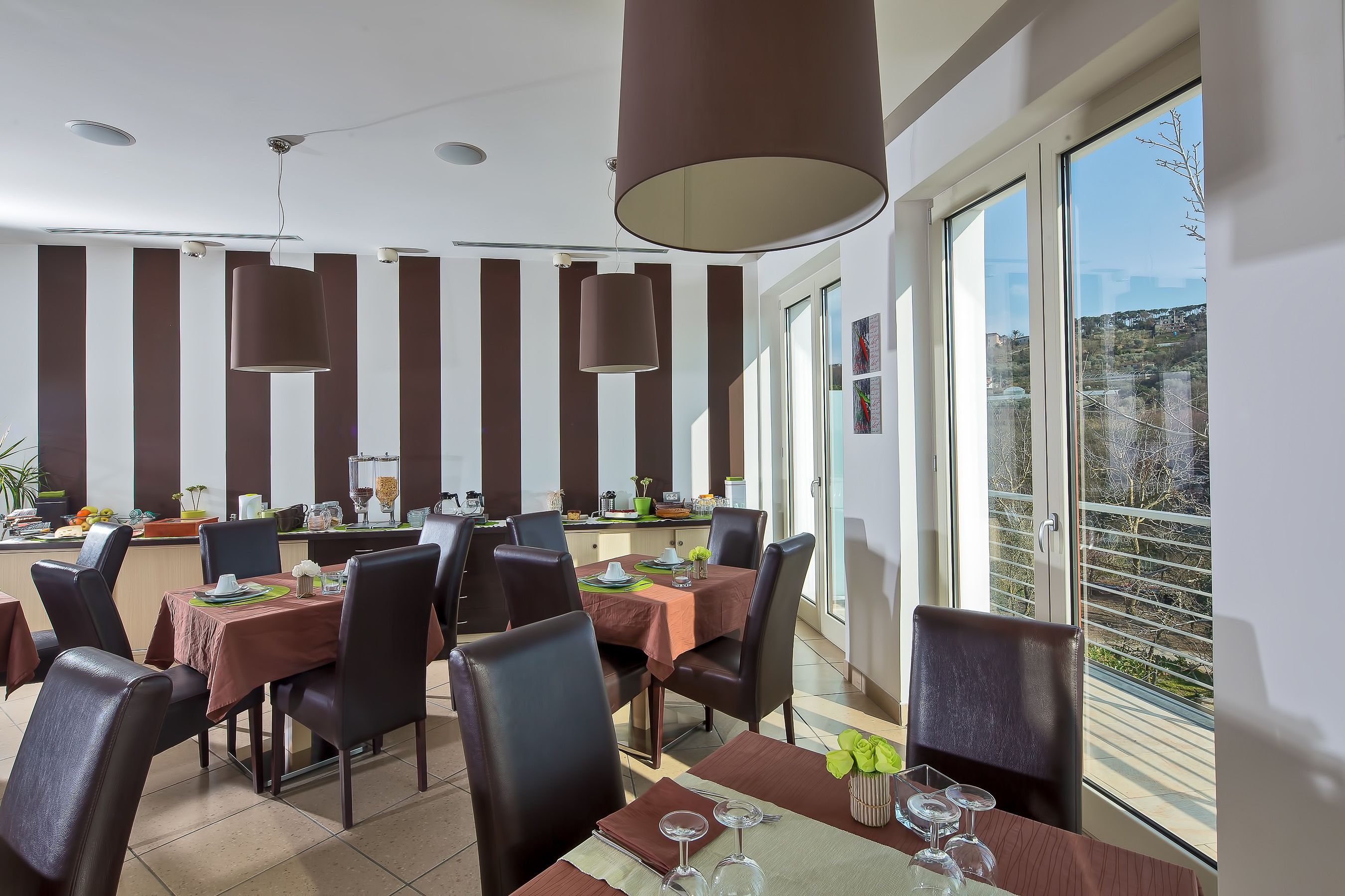 A dining area with tables set for four, large pendant lights, and a view of the outdoors through large windows.