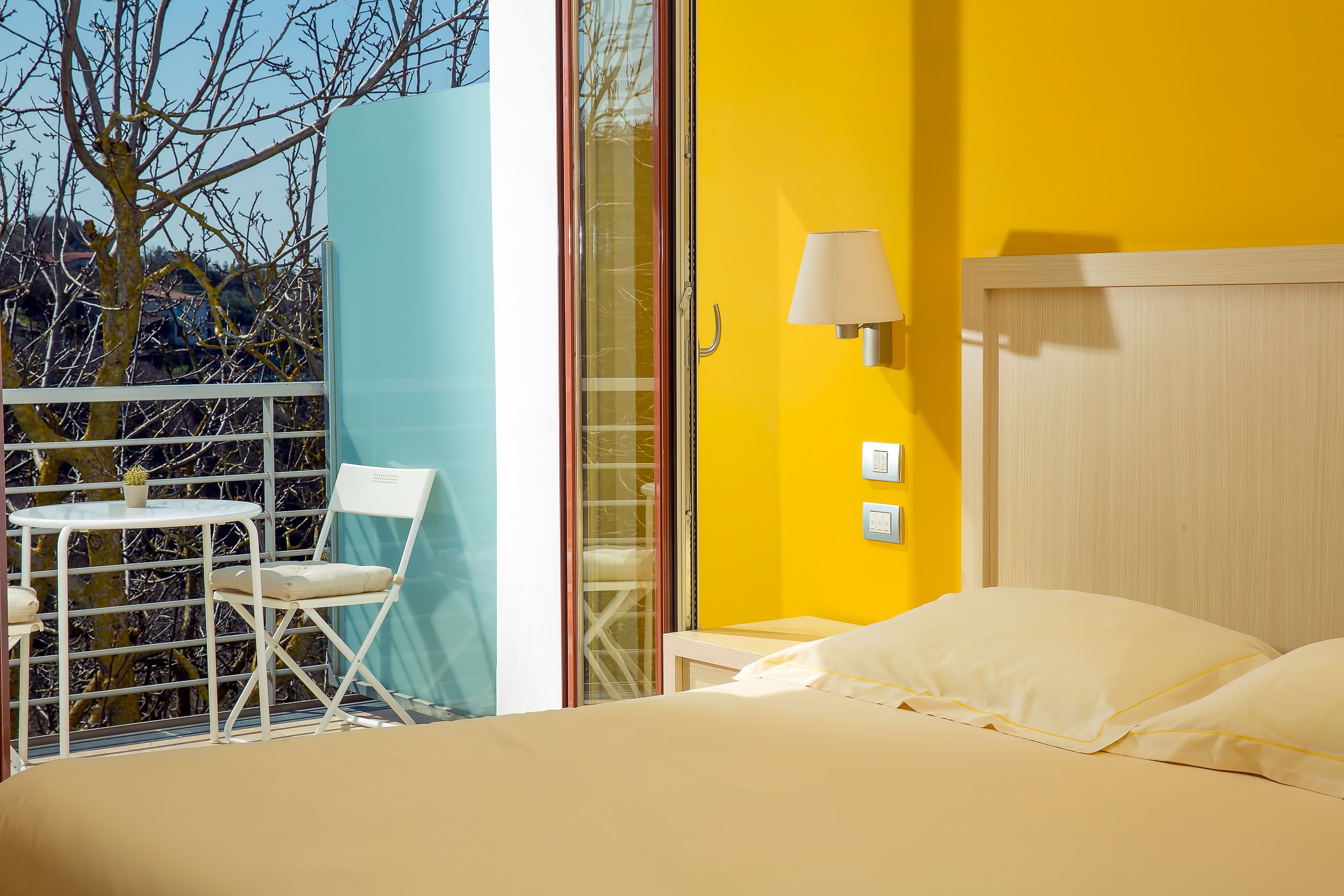 Ein Schlafzimmer mit Bett, Nachttisch, Lampe und Fenster mit Blick auf Bäume und ein Gebäude.