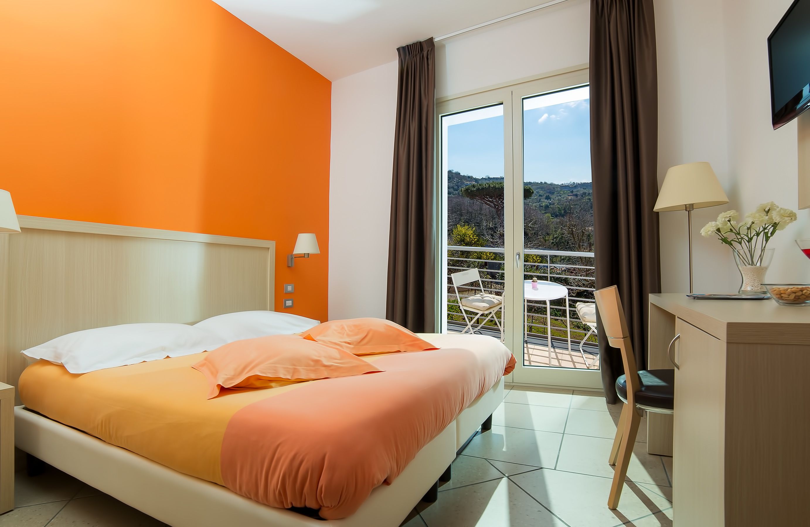 Habitación de hotel con cama, escritorio, silla, televisor y balcón con vistas a las colinas.